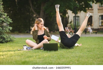 Caucasian Cute Couple Of College Students Messing Around On The Lawn In The Garden. Two Funny Students Being Silly, Having Fun In The University Park, Laptop, Coffee And Books Near Them.