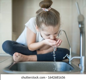 Caucasian Cute Girl Is Drinking From Water Tap Or Faucet In Kitchen At Home. Hydration. Lifestyle. Healthcare, Healthy Drink Concept. Fresh Clean Water. Thirsty Child. World Water Day