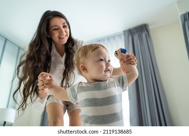 Caucasian Cute Little Baby Boy Child Learn To Walk With Mom In House. Happy Family, Mother Support Young Toddler Son Taking First Step Walk On Floor To Father To Develop Skill In Living Room At Home.