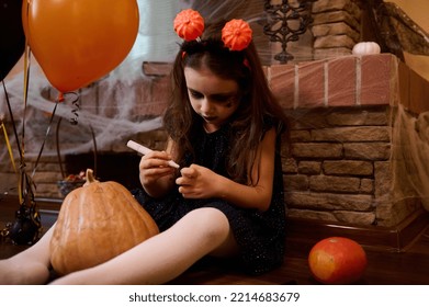 Caucasian cute little child  gothic girl in witch dress sitting by fireplace covered and spider web  orange   black air balloons  drawing horror face pumpkin  Halloween festival party