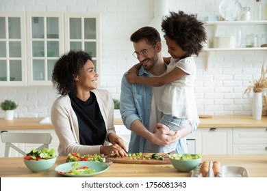 Caucasian dad holds on hands little mixed-race daughter while African wife preparing healthy vegetable salad family enjoy conversation in kitchen. Healthy home food, communication and cookery concept - Powered by Shutterstock