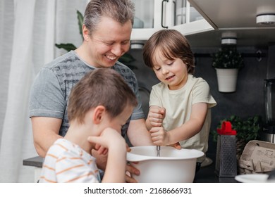 Caucasian dad with two children preparing dough for a pie or biscuit in the kitchen at home. One boy kneads the dough with a whisk in a bowl. Time with children