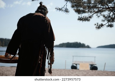 Caucasian dark-haired man in long clothes and with a tambourine goes to the lake. Shaman before the ceremony. Sandy beach. - Powered by Shutterstock