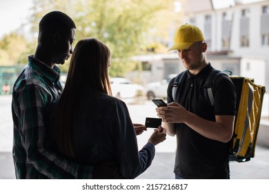 Caucasian delivery man delivering pizza or fast food. Young woman with husband at the door, pay by card for online order. Fast Home Delivery. Beautiful multiracial family wife and husband.