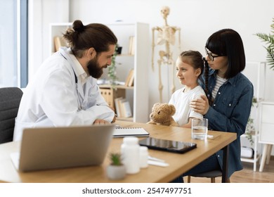 Caucasian doctor comforts young Caucasian girl while mother supports her during medical examination. Girl holds teddy bear, creating comforting atmosphere. - Powered by Shutterstock