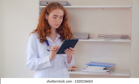 Caucasian Doctor With Red Curly Hair Reading Medical Records Using Digital Tablet. Candid Adult Woman Wearing In White Lab Coat And Phonendoscope Standing In Hospital.