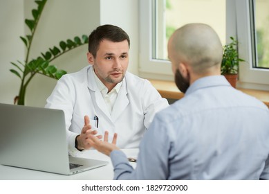 A caucasian doctor in a white lab coat is sitting at a desk and describing the treatment of a bald male patient in a hospital. A man with a beard at an appointment in a doctor's office. - Shutterstock ID 1829720900