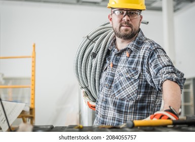 Caucasian Electrician Contractor Worker in His 30s with Metal Electric Cables Conduit Pipe on His Shoulder. Construction Site Theme.