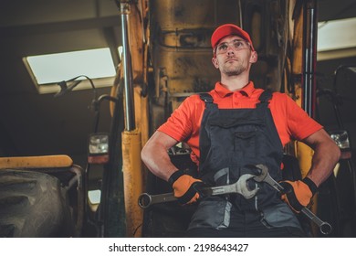 Caucasian Engineer Sitting In Front Of Heavy Duty Vehicle With Wrenches In His Hands Taking A Short Break During Work. Industrial Machinery Repair Shop.