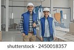 Caucasian engineers with safety helmets cheerfully smiling while looking at camera. Workers holding tablet device and laptop. Enjoying working at industrial facility. Around people located crates.