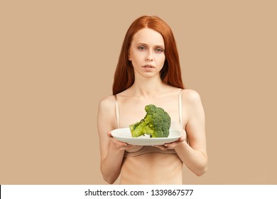 Caucasian exhausted redhead woman with undereye circles holding fresh broccoli looking at camera with tired depressed expression