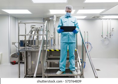 caucasian factory worker in blue lab uniform, blue gloves and blue facemask stand on chromed metal ladder, look in tablet, steel tank on background - Shutterstock ID 573764839