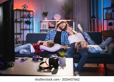 Caucasian Family Of Four Fall Asleep While Watching Bored Movie On TV. Young Parents With Two Kids Sitting On Couch With Closed Eyes, Sleeping In Front Of Working TV Set