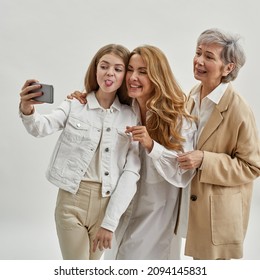 Caucasian family of grandmother, mother and granddaughter taking selfie on smartphone. Age and generation concept. Three female generations. Young girl sticking out tongue. White background in studio - Powered by Shutterstock