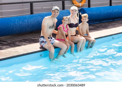 Caucasian Family Having Fun By Their Swimming Pool. Happy Young Family Splashing Water With Hands And Legs While Sitting On Edge Of Swimming Pool. Kids With Parents Playing Outdoors