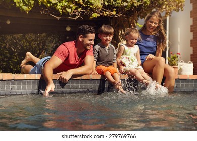 Caucasian family having fun by their swimming pool. Happy young family splashing water with hands and legs while sitting on edge of swimming pool. Kids with parents playing outdoors. - Powered by Shutterstock