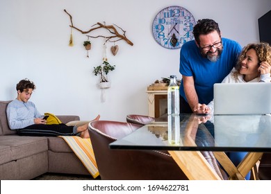 Caucasian Family At Home Lifestyle With Technology - Couple Husband And Wife Use Laptop Computer On The Table And Teenager Son With Phone On The Couch - Quarantine Lockdown Concept