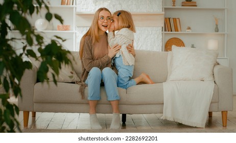 Caucasian family at living room. Woman mother babysitter listen secret from little daughter kid baby child girl whisper talking gossip in mommy ear share secrecy information rumor sitting at couch - Powered by Shutterstock