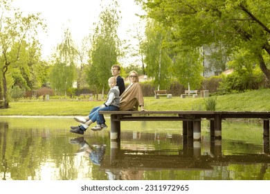 Caucasian Family Mother, Father And Son Sit On Wooden Dock Enjoying Lake View In Summer Time. Family Support And Care. Parenthood, Family Leisure Time. Children's Day. Horizontal Plane. - Powered by Shutterstock
