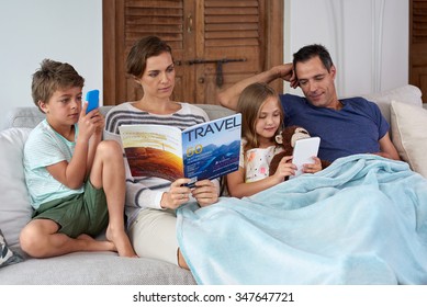 Caucasian Family Relaxing At Home Mom Reading Magazine Kids Playing Games On Tablet Device And Mobile Cell Phone