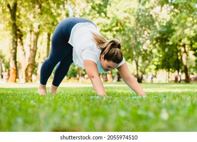 Caucasian fat plump body positive plus size standing in yoga position downward facing dog on fitness mat, slimming, stretching outdoors in park