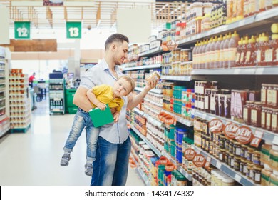 Caucasian Father Dad In Grocery Store Shopping Buying Food And Holding Carrying Baby Son Under Arm. Man Parent With Toddler Kid Choosing Meal For Dinner Lunch. Difficult Parenting Authentic Moment.