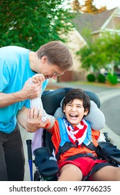 Caucasian father helping disabled ten year old son in wheelchair adjust orthotic arm guards outdoors. Child has cerebral palsy