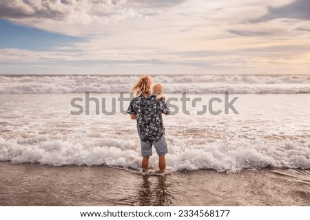 Caucasian father holding child in his arms. Happy childhood. Dad and baby son spending summer vacation on the beach. Horizon line. Ocean, waves, cloudy sky. View from back. Seminyak, Bali, Indonesia