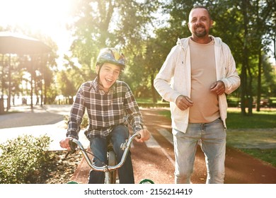Caucasian father run while his son with cerebral palsy ride bicycle in park. Smiling man and boy looking at camera and spending time together. Family relationship. Disability care and rehabilitation