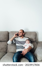 Caucasian Father Sitting On Couch With Baby In His Arms.The Man Is Talking On A Cell Phone While Hugging His Little Daughter.