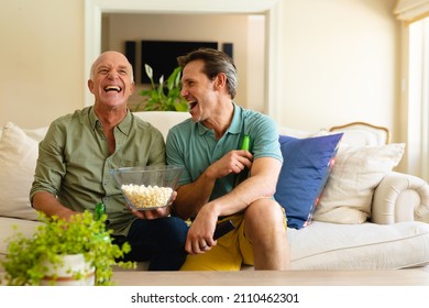 Caucasian father and son with beer and popcorn watching tv sitting together on couch at home. sports and entertainment concept, unaltered.