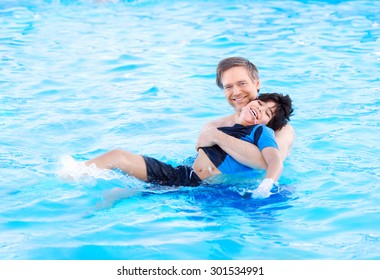 Caucasian Father Swimming In Pool With Biracial Disabled Son In His Arms. Child Has Cerebral Palsy.