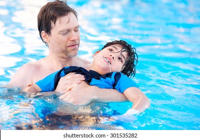 Caucasian Father Swimming In Pool With Biracial Disabled Son In His Arms. Child Has Cerebral Palsy.