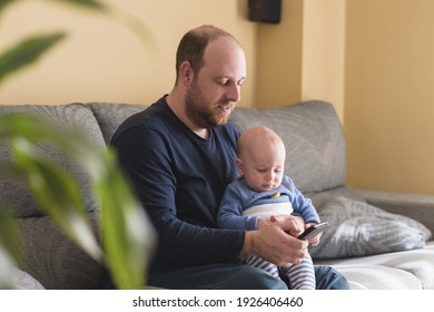 Caucasian Father Talking On Mobile Phone To Work Online With His Newborn Baby. Single Dad Taking Care Of His Son. Happy Family And Work At Home.