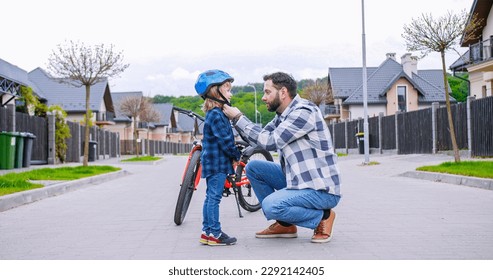 Caucasian father teaching small cute son riding a bike. Dad putting on helmet on head of little boy. Outdoor. Learning how to ride bicycle. - Powered by Shutterstock