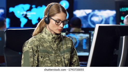 Caucasian female army officer in glasses and headset talking, working on computer in analytic secret base in military. Woman militarian official typing in dark office. Digital support Warfare operator