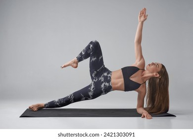 Caucasian female athlete in dark activewear doing side plank with bent leg in studio. Side view of concentrated sportswoman practicing variation of side plank asana, isolated on grey. Yoga concept.