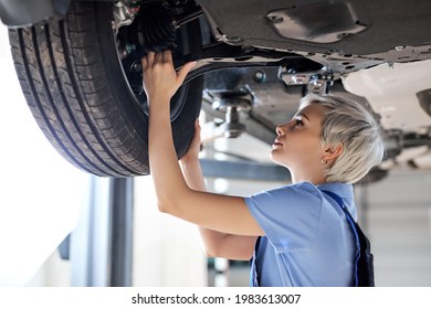Caucasian female auto mechanic changing wheel tire in car in garage, side view. Beautiful young lady in overalls is concentrated on work, carefully adjusting repairing