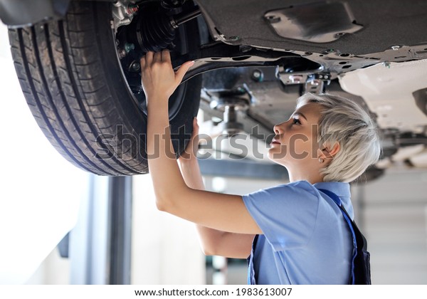 Caucasian female auto\
mechanic changing wheel tire in car in garage, side view. Beautiful\
young lady in overalls is concentrated on work, carefully adjusting\
repairing