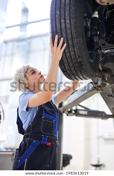 Caucasian female auto\
mechanic changing wheel tire in car in garage, side view. Beautiful\
young lady in overalls is concentrated on work, carefully adjusting\
repairing