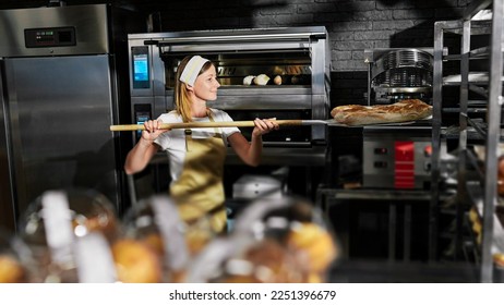 Caucasian female baker in apron and hat taking out just-baked baguettes from oven and putting on shelf in kitchen of bakehouse. Woman working in bakery and baking fresh bread. Cooking profession. - Shutterstock ID 2251396679