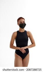 Caucasian female ballet dancer in medical mask stand and look away. Concept of health protection during Coronavirus pandemic. Girl with red hair and wear leotard. Woman on white background in studio