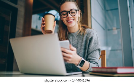 Caucasian female blogger in trendy spectacles smiling indoors while reading received email with funny video file connected to 4g wireless in coffee shop, happy woman with takeaway cup chatting via app