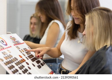 Caucasian Female Client And Asian Hairdresser, Hair Stylist In Beauty Salon Choosing Hair Color For Coloring, Dyeing On Palette In Catalog. Soft Focus