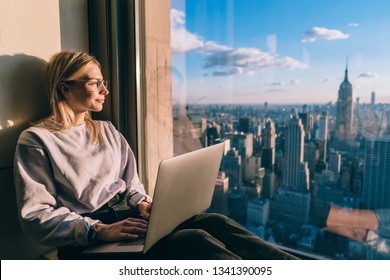 Caucasian female digital nomad sitting with laptop computer on window sill of modern skyscraper building coworking. Young woman freelancer enjoying downtown cityscape from hotel room during vacations - Powered by Shutterstock