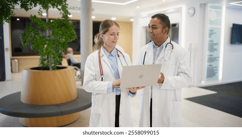 Caucasian Female Doctor Briefing an Indian Male Physician on Patient Data on a Laptop Computer. Bright Hospital Hall Busy with Activity as Medical Professionals Walk Towards Their Departments - Powered by Shutterstock