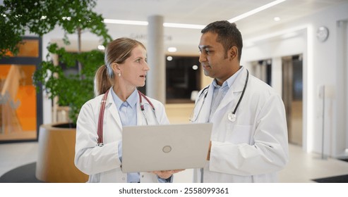 Caucasian Female Doctor Briefing an Indian Male Physician on Patient Data on a Laptop Computer. Bright Hospital Hall Busy with Activity as Medical Professionals Walk Towards Their Departments - Powered by Shutterstock