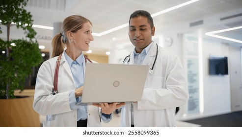 Caucasian Female Doctor Briefing an Indian Male Physician on Patient Data on a Laptop Computer. Bright Hospital Hall Busy with Activity as Medical Professionals Walk Towards Their Departments - Powered by Shutterstock