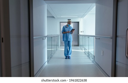 Caucasian Female Doctor In Hospital Wearing Face Mask And Scrubs Using Tablet. Medical Professional At Work And Health Services During Coronavirus Covid 19 Pandemic.