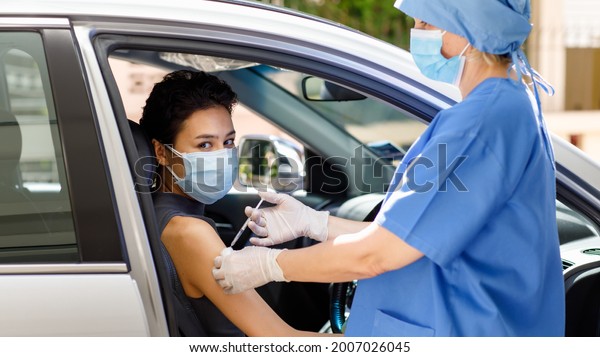 Caucasian female doctor from public health in blue
hospital uniform and face mask stand hold vaccine syringe needle
injection on woman patient shoulder in drive through car
vaccination queue.